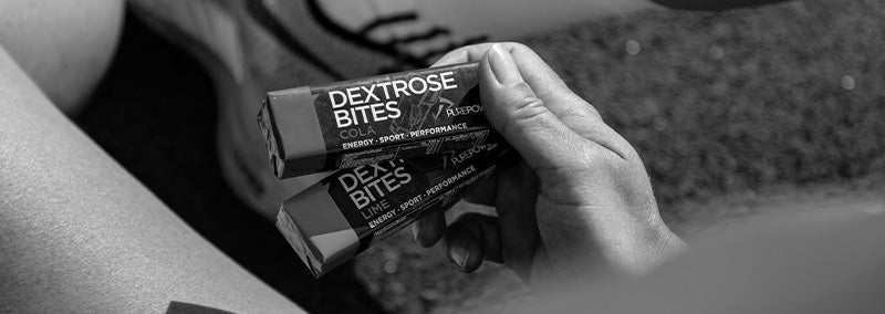 Close-up av en hånd som holder to Dextrose Bites energiprodukter i et treningsmiljø – illustrerer rask karbohydrat som pre-start-snack eller supplement til frokost før maraton.